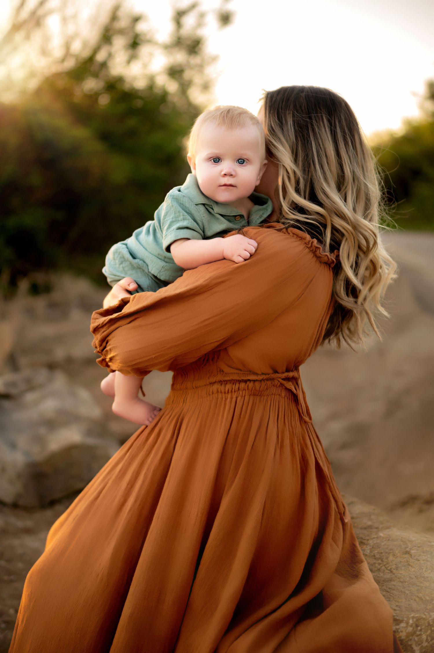 A tender moment captured between parent and child in a flowing rust-colored dress at sunset.