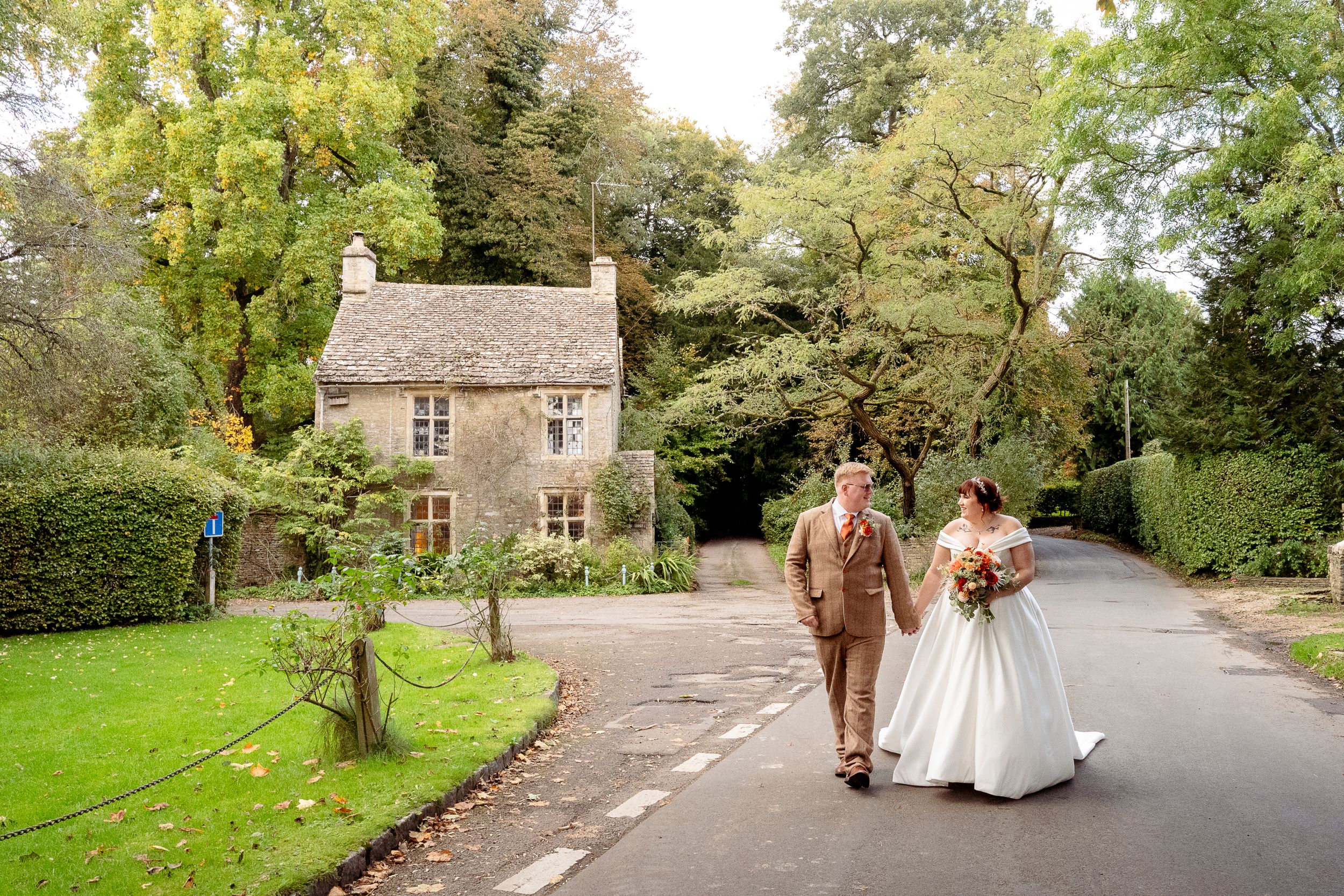 Newlyweds walking down country lane past quaint stone cottage.