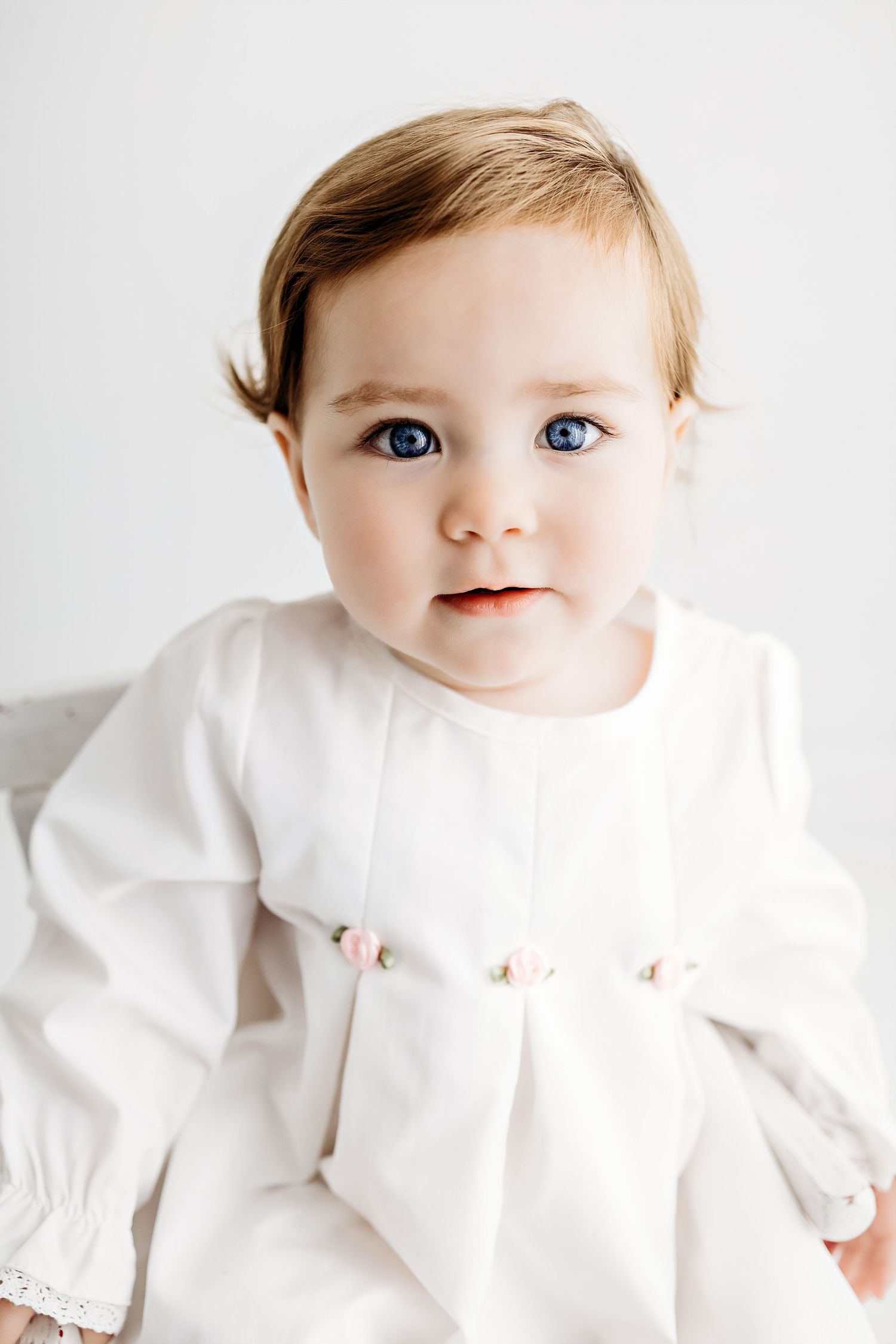 Baby wearing an elegant white dress with lace details against light background.