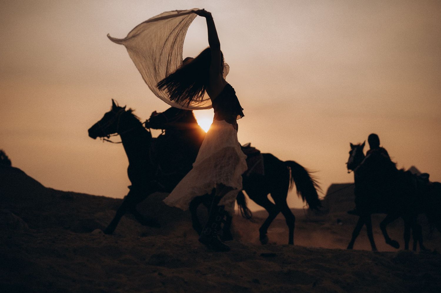 Silhouetted riders on horseback with flowing fabric banner against dramatic sunset sky in desert landscape.