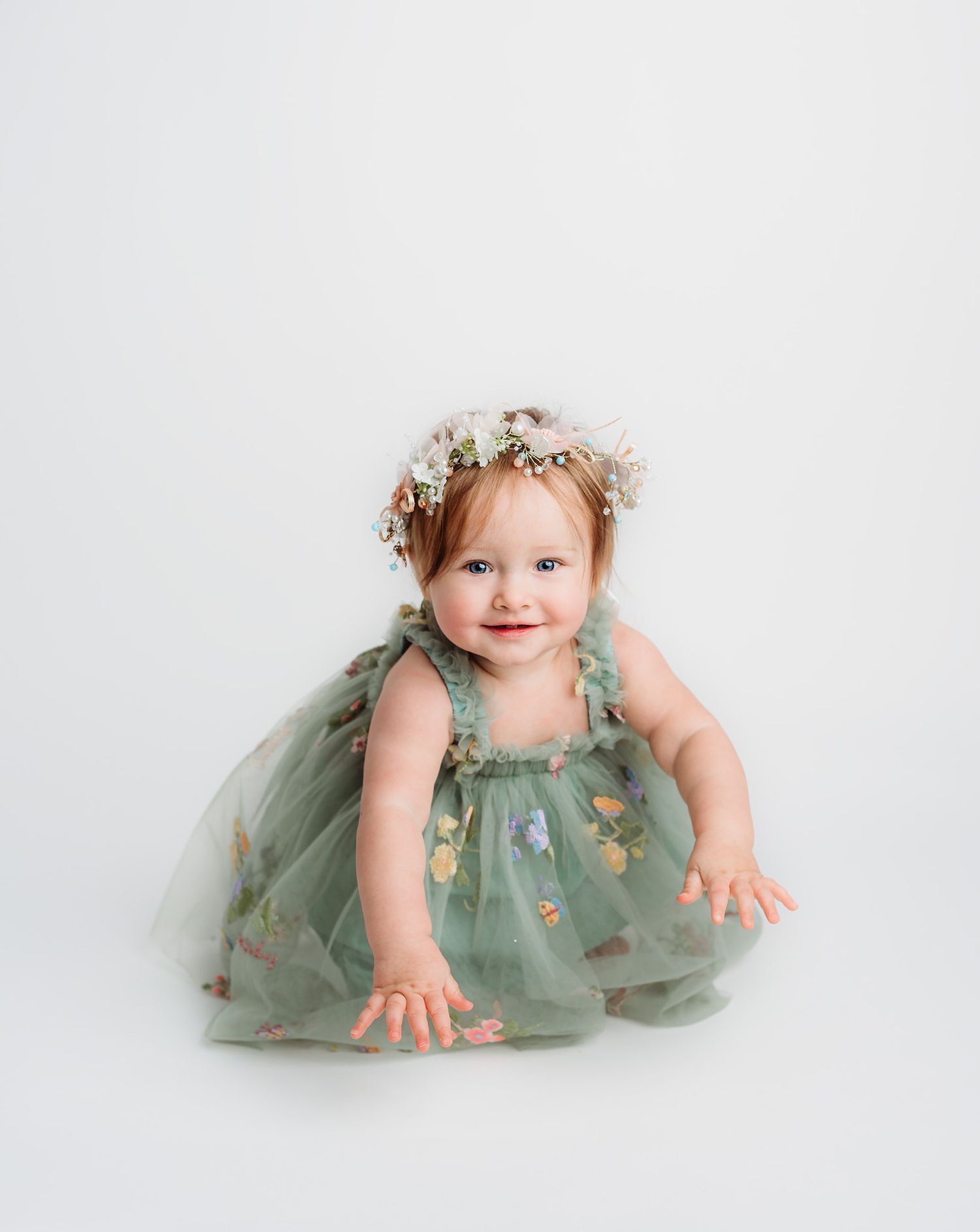 Smiling baby girl in green floral tulle dress and flower crown during studio portrait session.