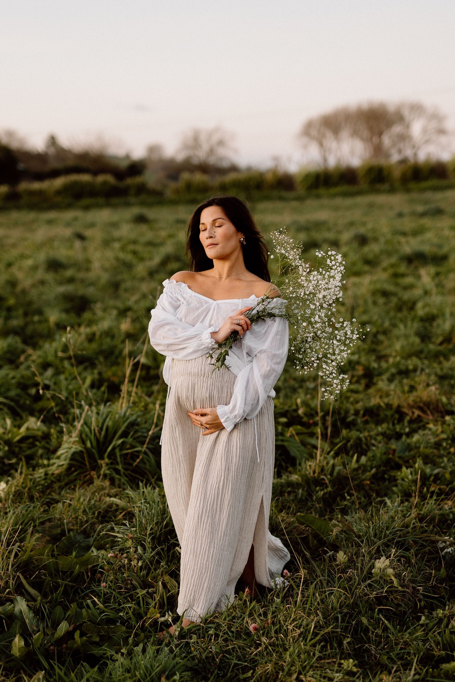 Ethereal maternity photoshoot in a grassy field with flowing white dress and delicate flowers at golden hour.