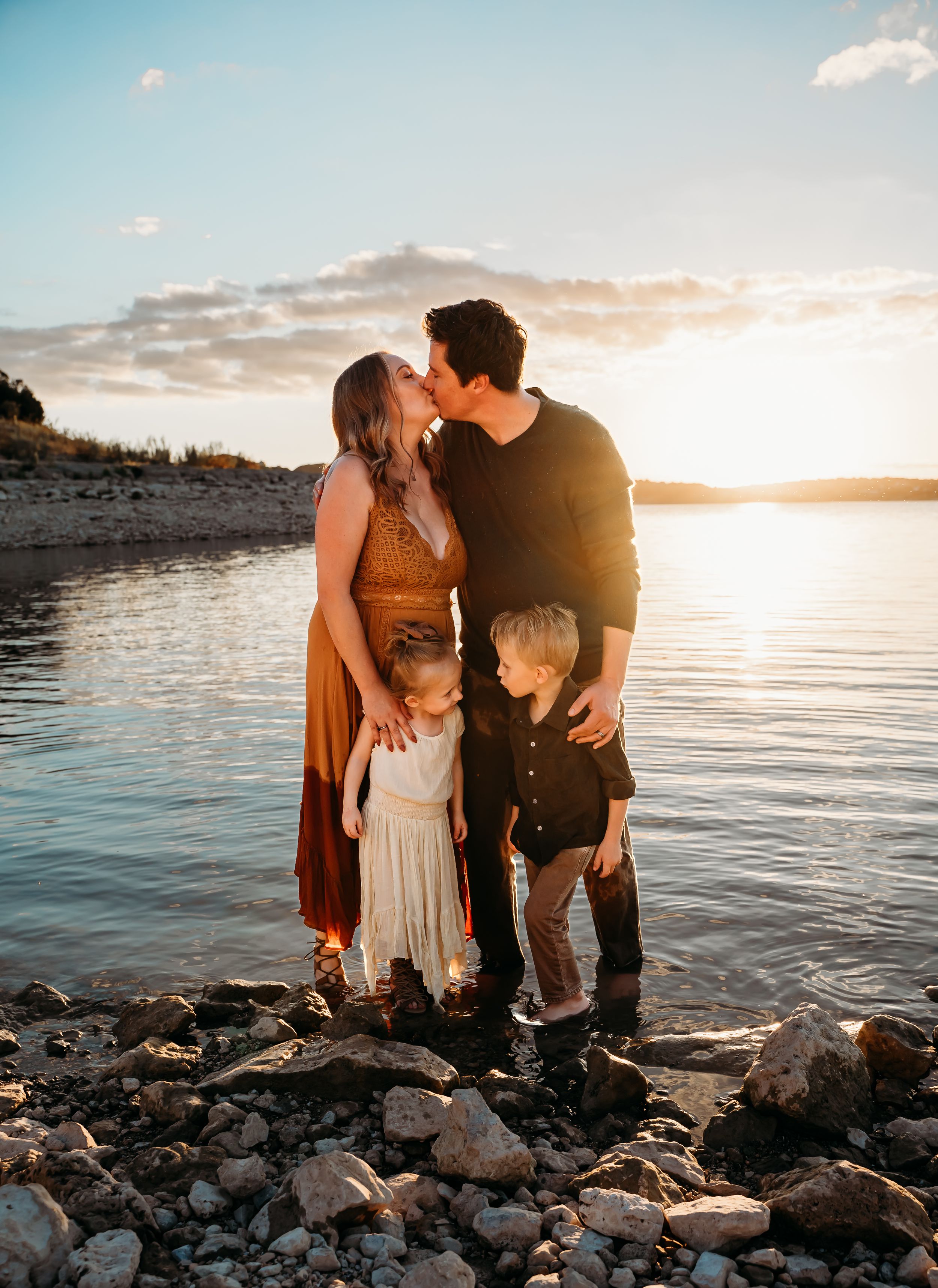 Family standing together on rocky shoreline at sunset with water backdrop.