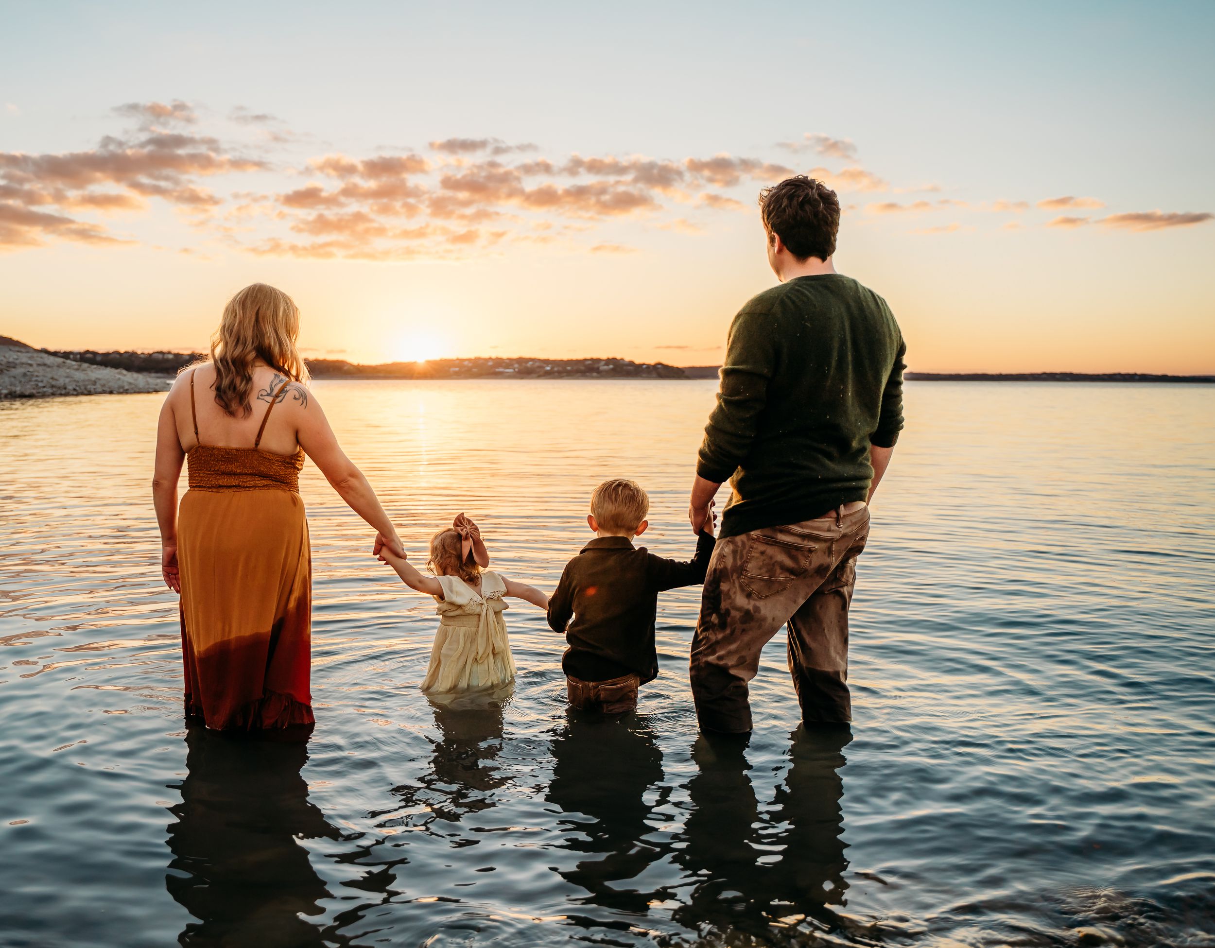 Family standing together in lake water during golden sunset creating silhouette against orange sky.