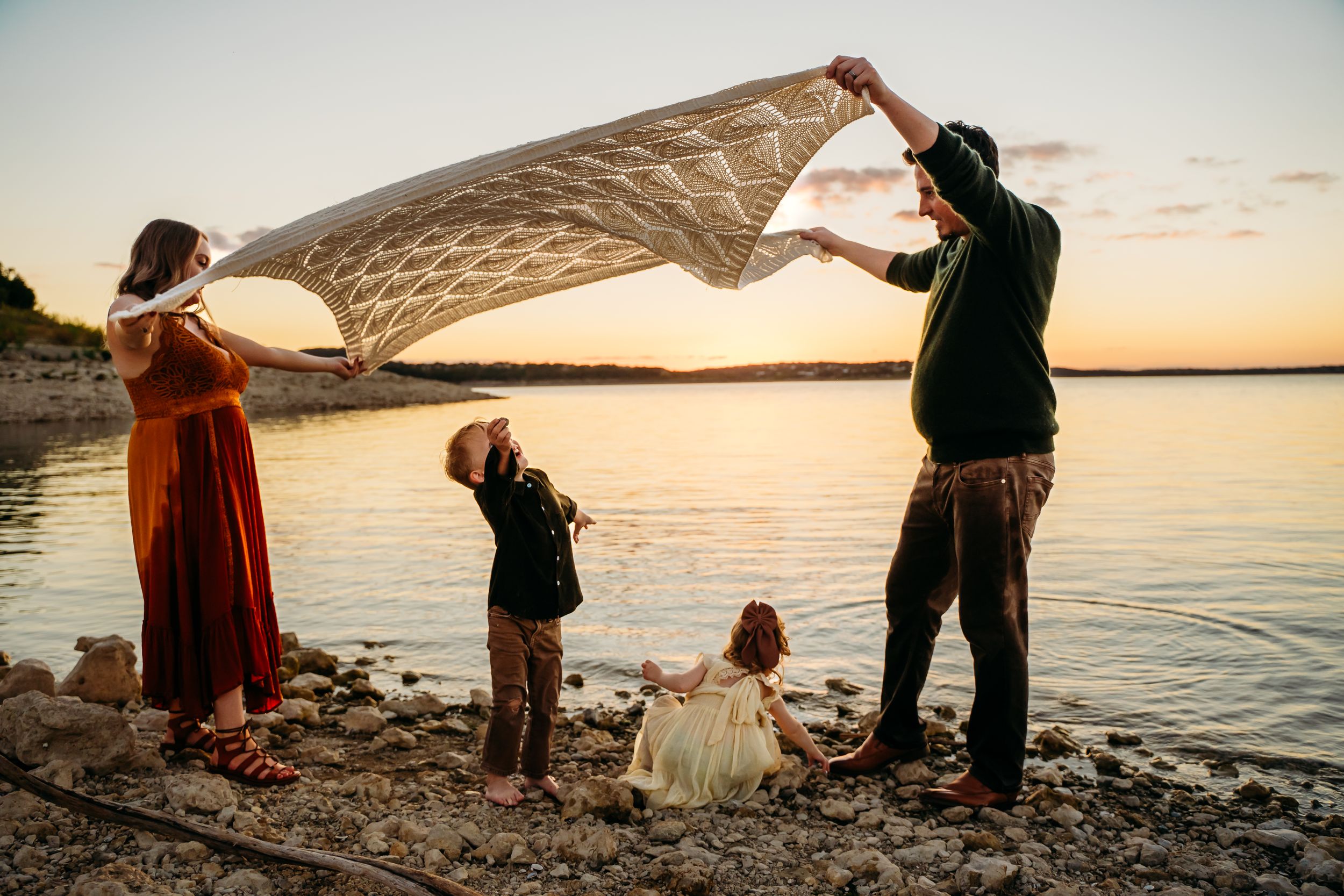 Family holds blanket up together on beach during golden sunset.