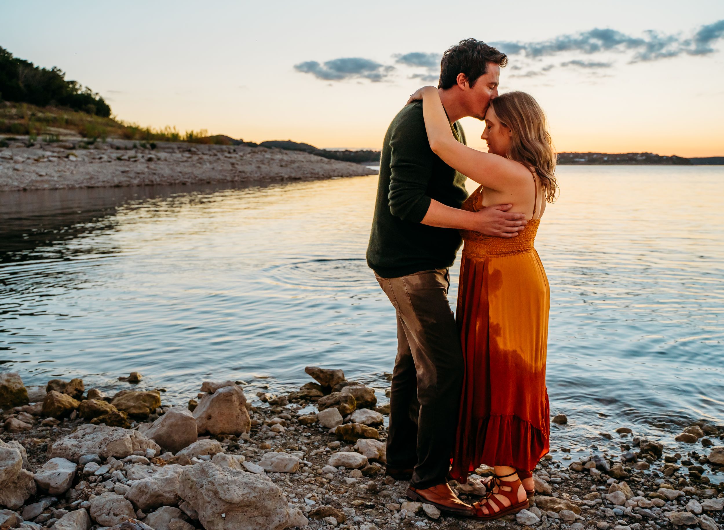 Couple in romantic embrace on rocky beach shore during sunset.