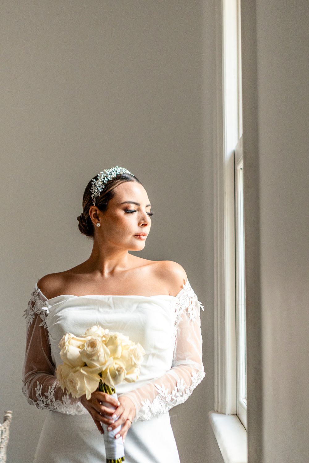 A bride in an elegant off-shoulder white gown with lace sleeves holds a cream bouquet while standing by a window.