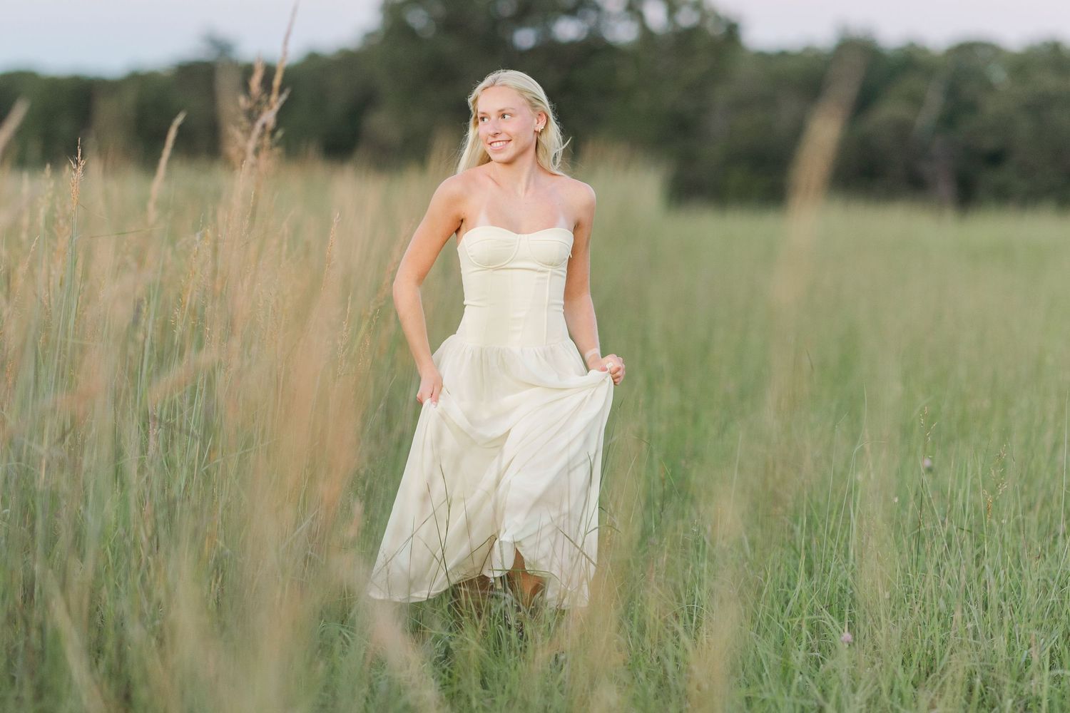 Senior girl in a white flowing dress sits on a wooden dock by the water on a bright sunny day.