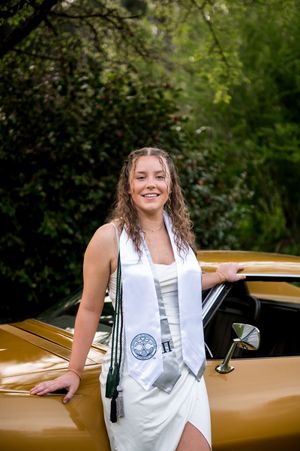 A white suit-clad figure poses playfully against a shiny gold classic car in an outdoor setting.