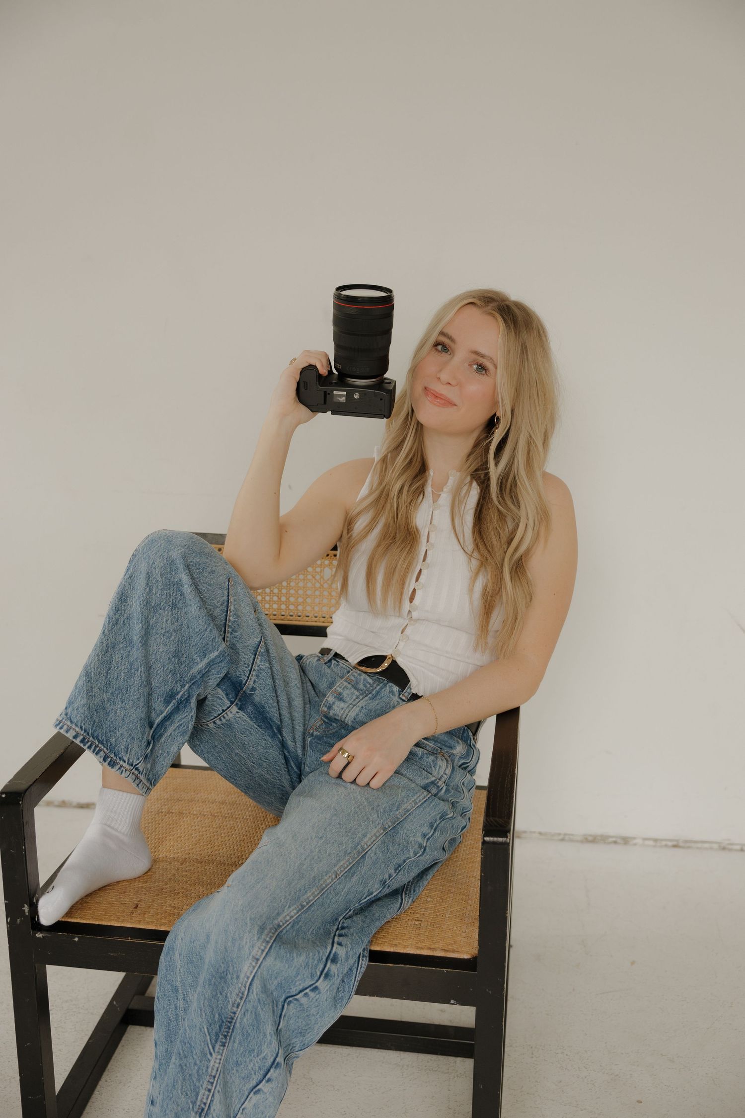 Blonde female photographer sitting in a wicker chair holding a camera in a bright white studio.