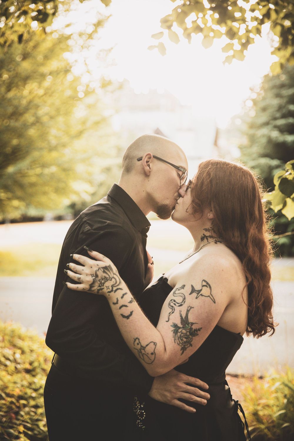 Romantic couple sharing intimate kisses in golden sunlit garden setting with visible tattoos and natural foliage.