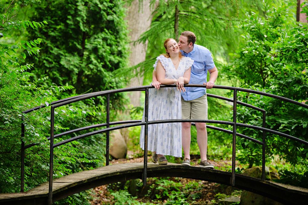 An engaged couple shares a romantic moment on a rustic wooden bridge surrounded by lush green forest at Canton Garden Center in Ohio