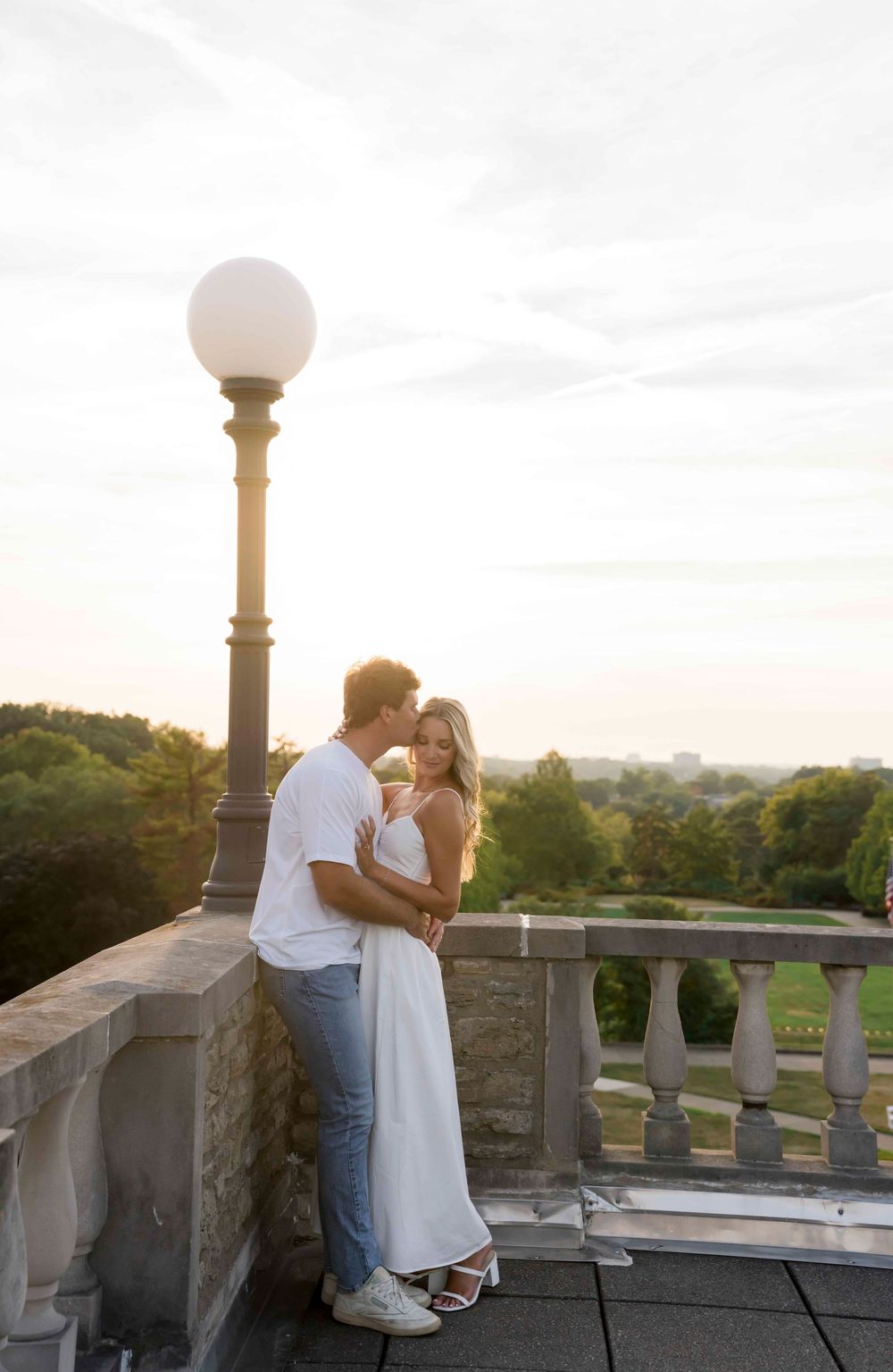 Couple sharing an intimate moment at sunset on a stone balcony with ornate railing and glowing lamp post.