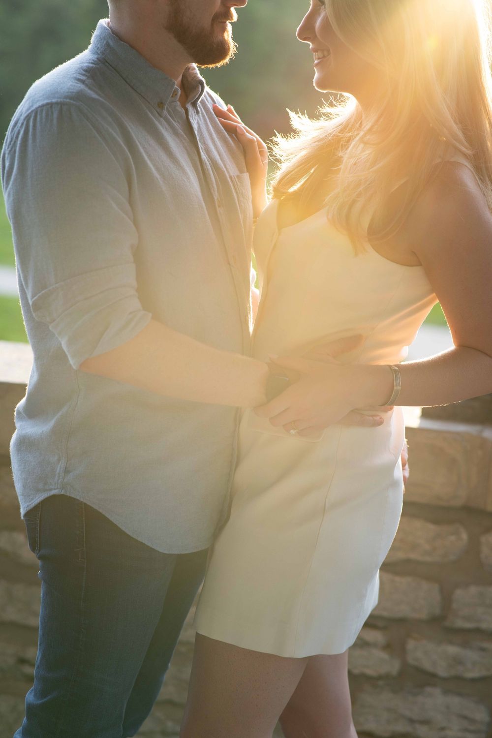 Couple embracing in warm golden sunlight, man in gray shirt and woman in white dress sharing a romantic moment outdoors.