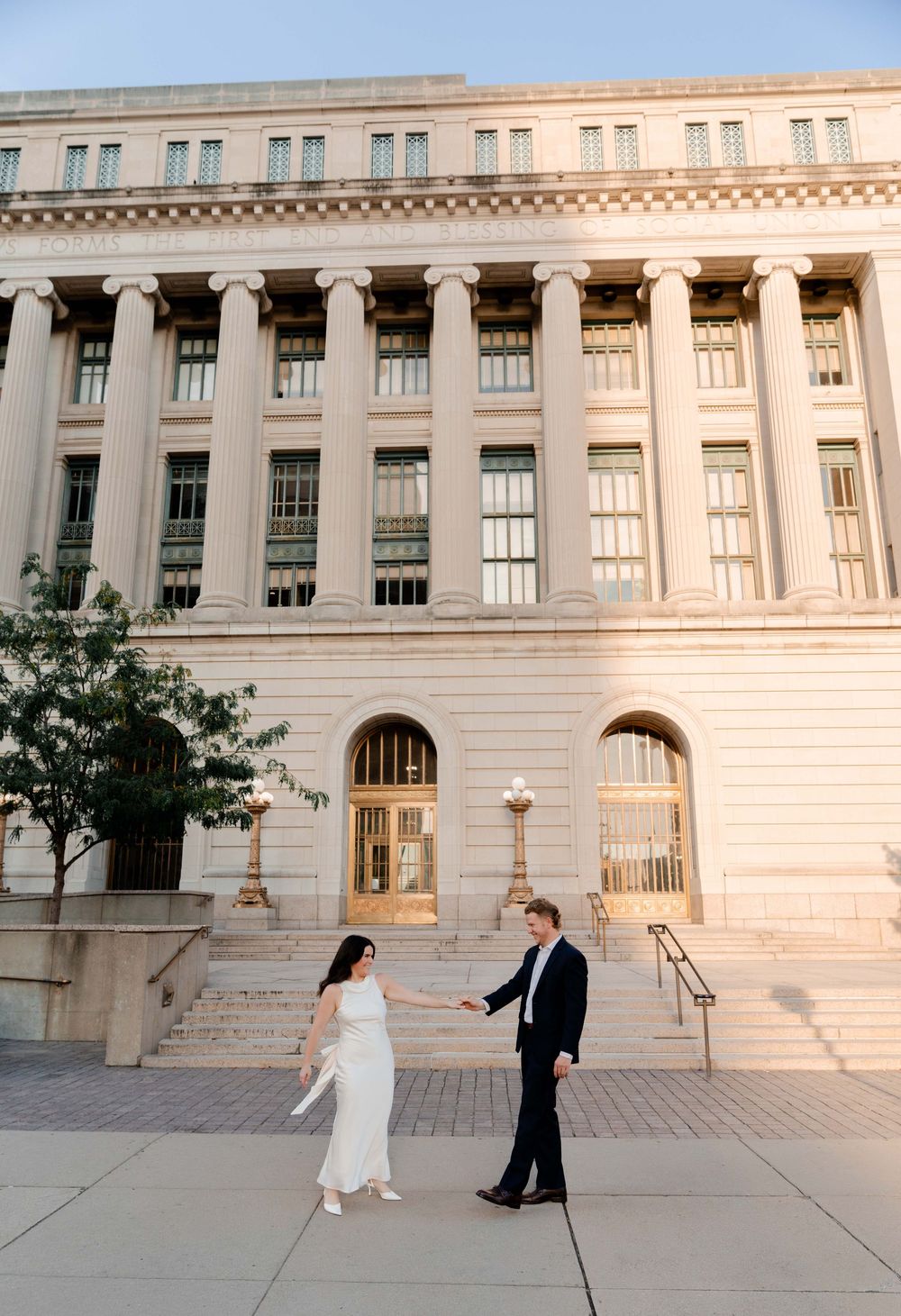 Couple in wedding attire holding hands outside a grand neoclassical building with tall columns at golden hour.
