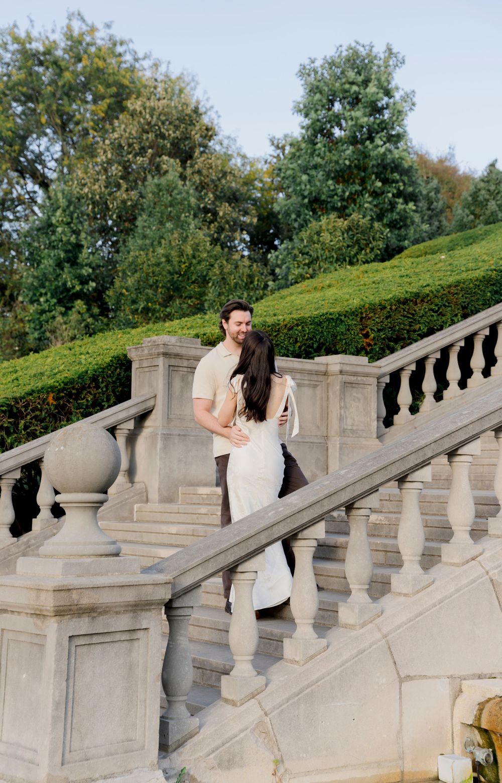 A couple shares a romantic moment on elegant stone steps surrounded by lush greenery and ornate balusters.