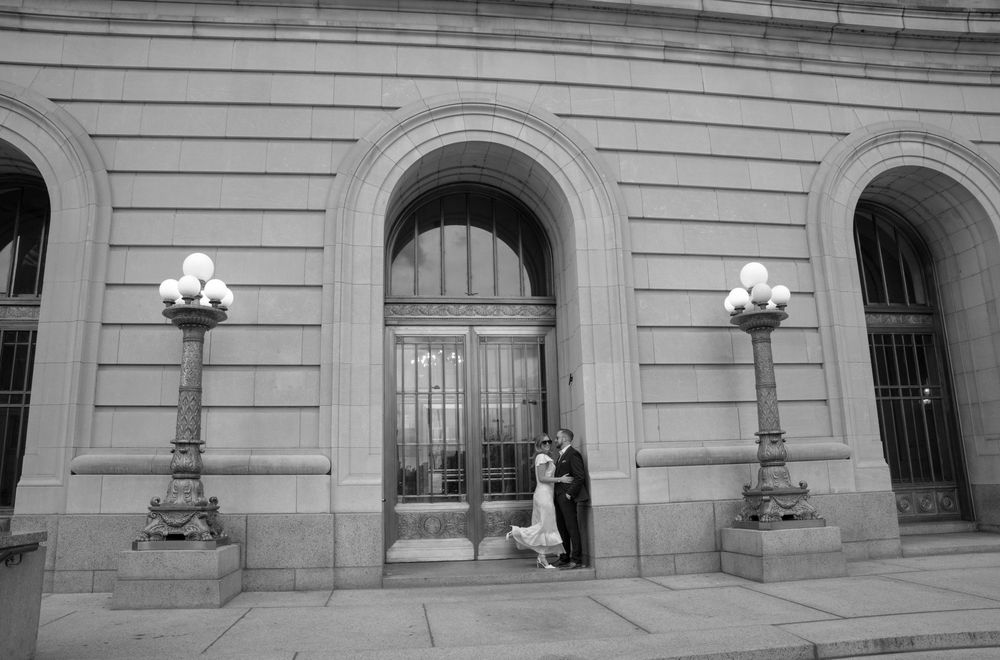 Black and white photo of a bride and groom posing under grand arched entrance of historic stone building.
