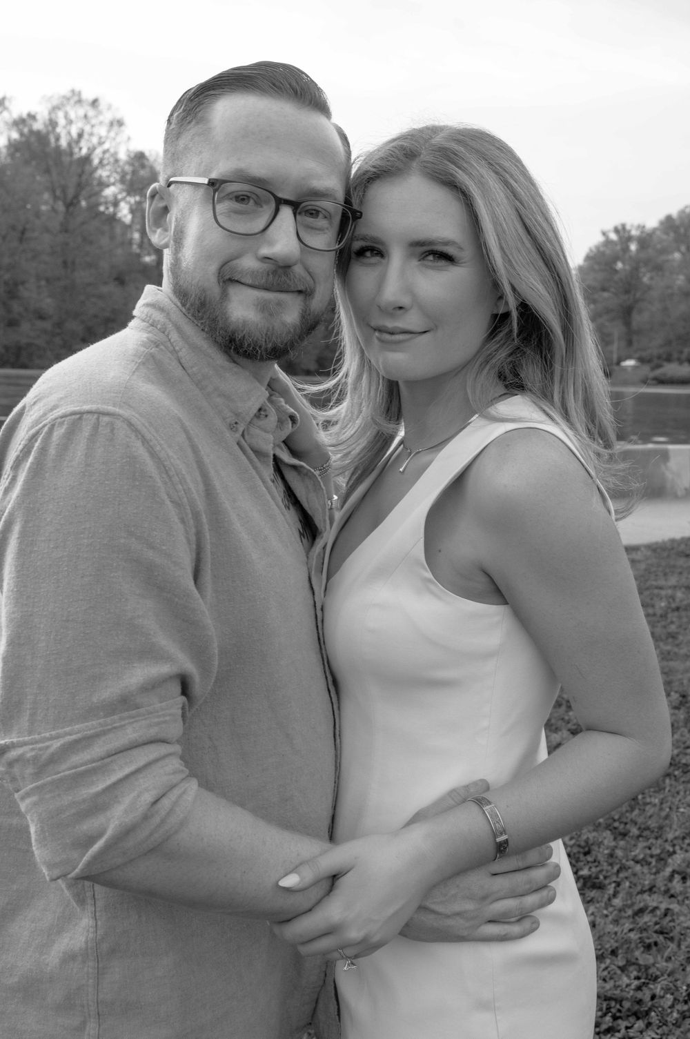 Black and white portrait of a smiling couple embracing outdoors, man wearing glasses and woman in white dress.