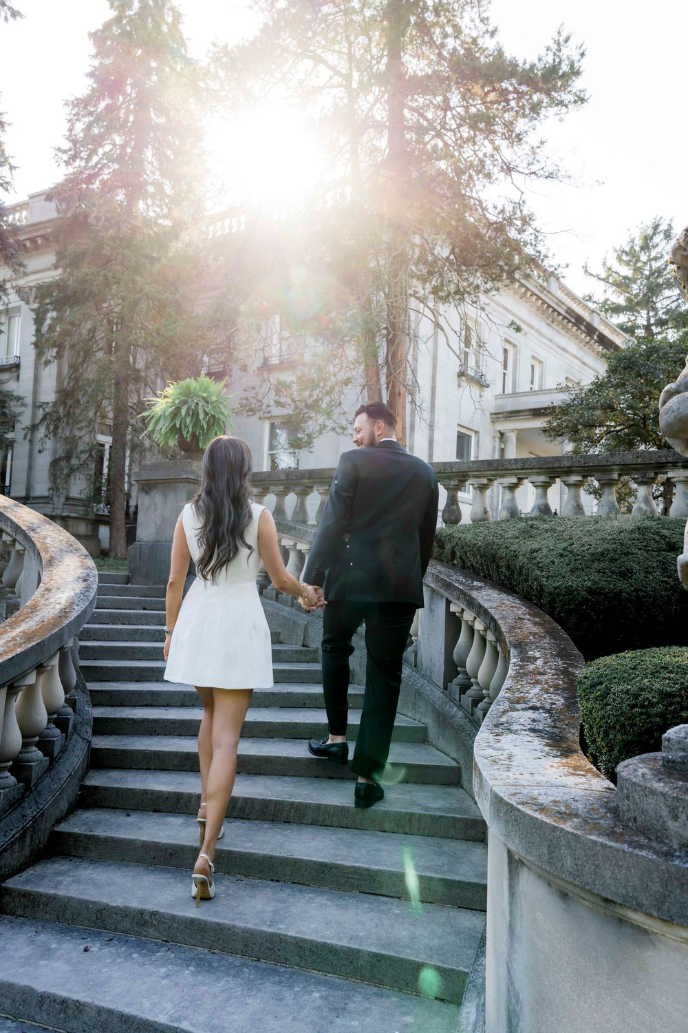 A couple walking up outdoor stone steps with sunlight flare in a garden setting at sunset.