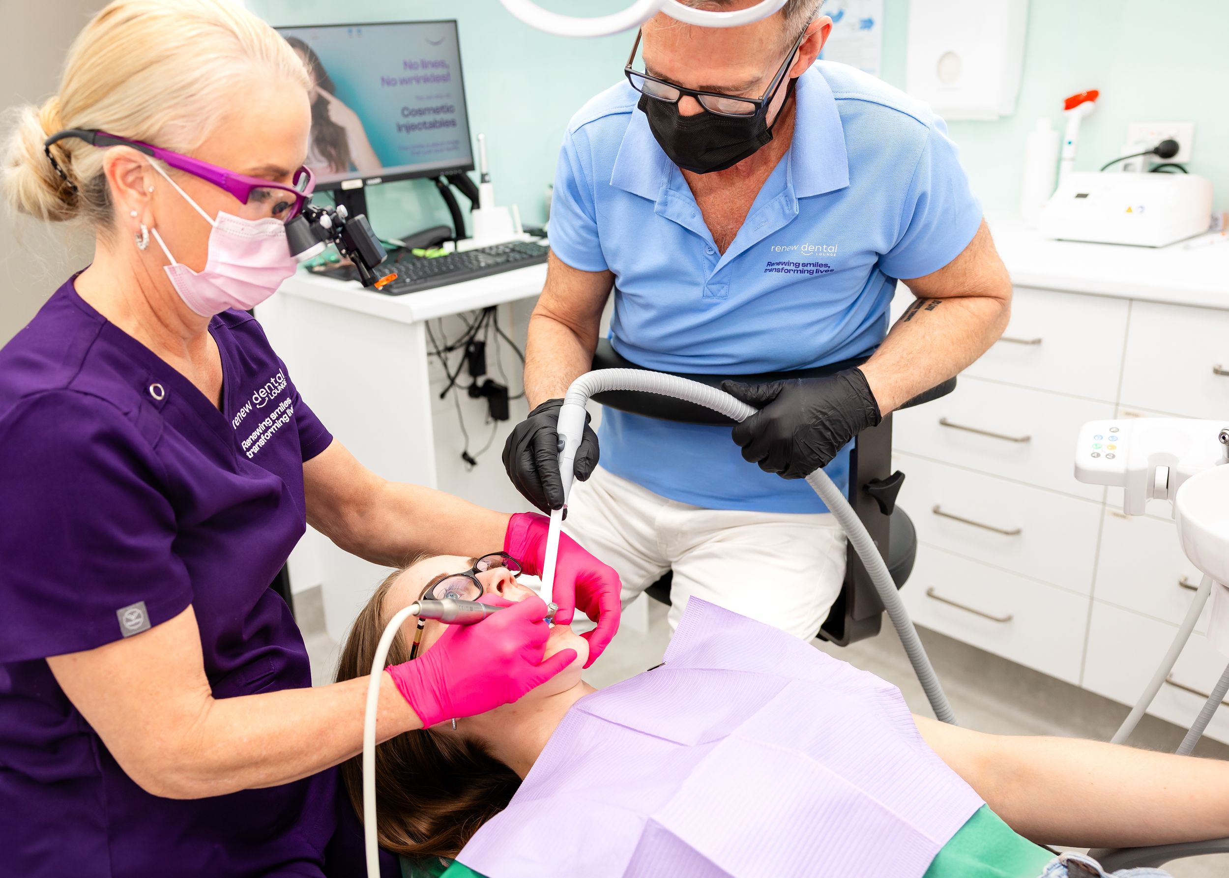 Healthcare professional in scrubs performing dental examination while patient reclines in dental chair.