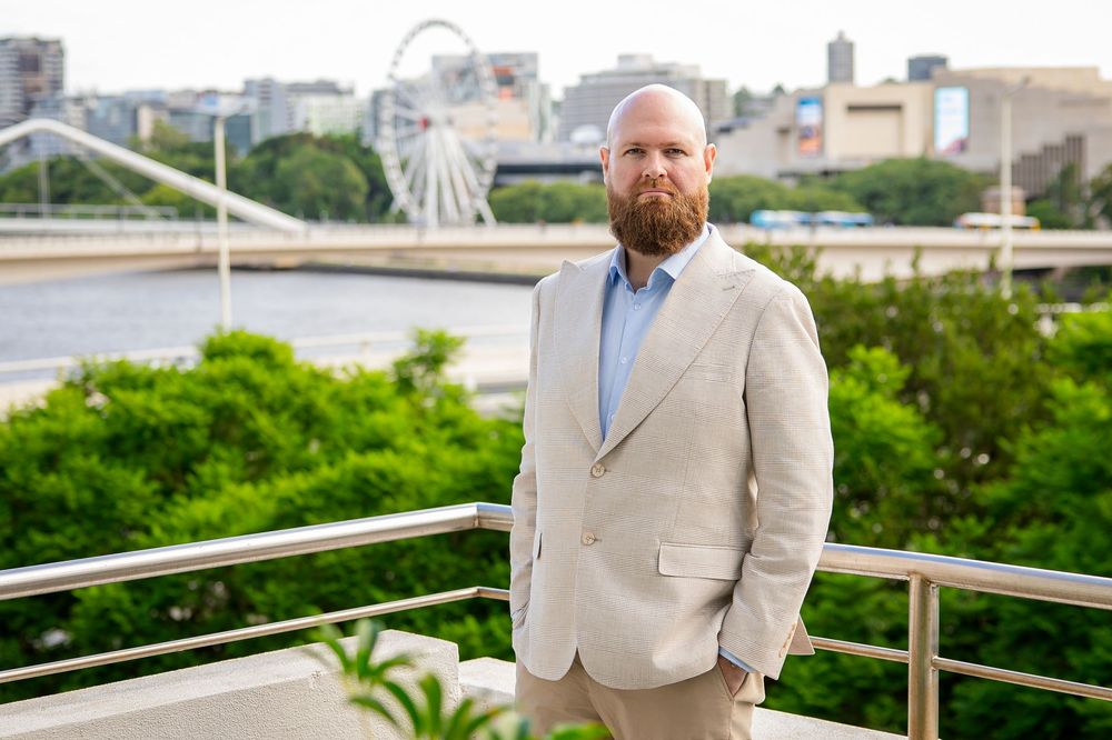 A groom in a light beige suit poses for wedding photos on an outdoor terrace with greenery in the background.
