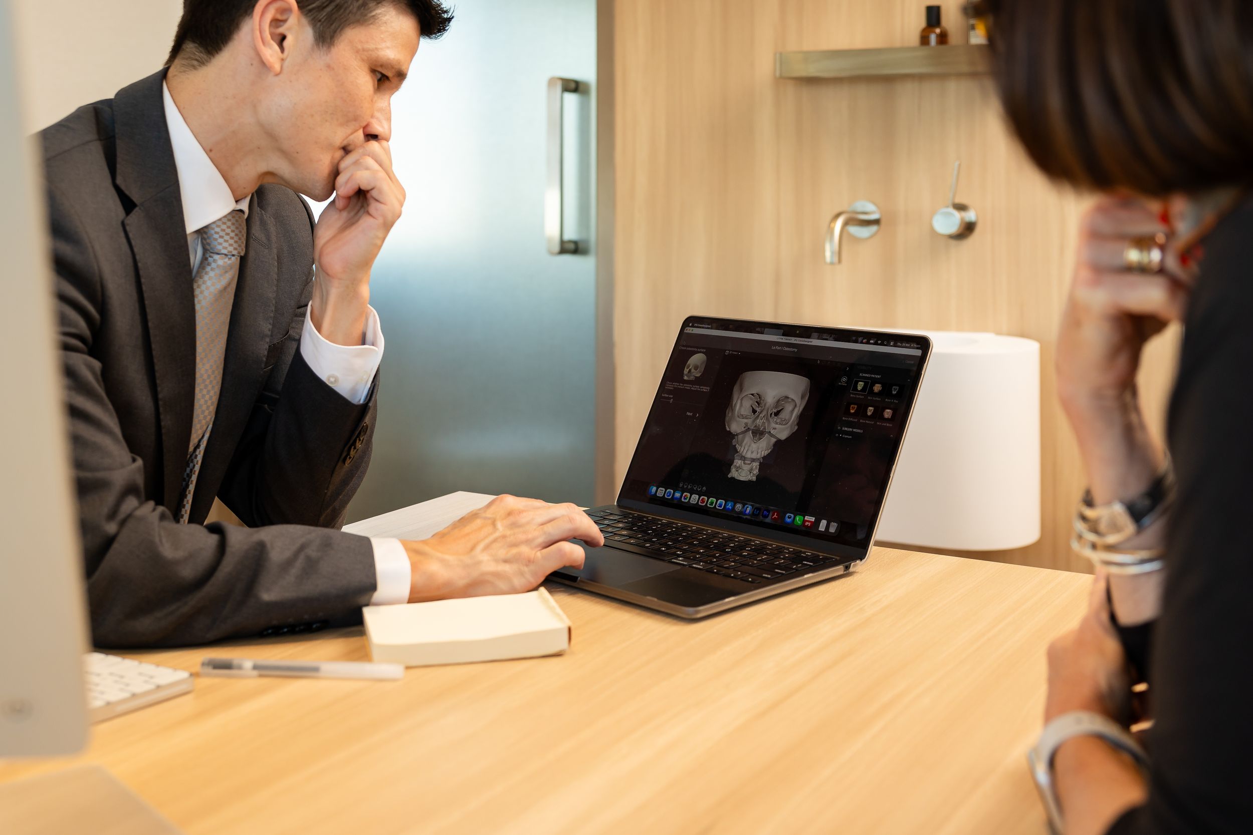 A male doctor in a suit reviews a 3D facial scan on a laptop during a patient consultation.