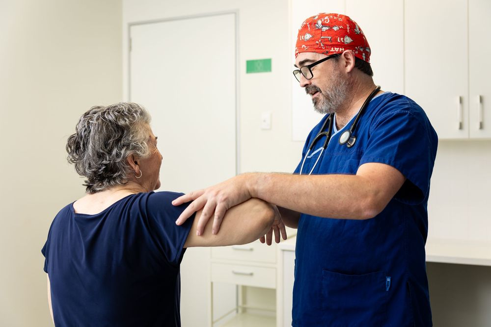 Healthcare provider in blue scrubs examining a patient's shoulder in a medical examination room.