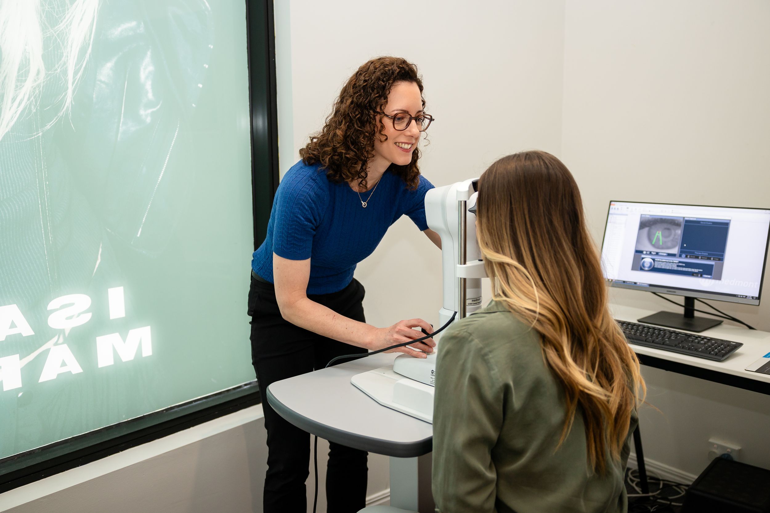 Two people having a discussion in an office setting with a computer monitor and projected display visible in the background.