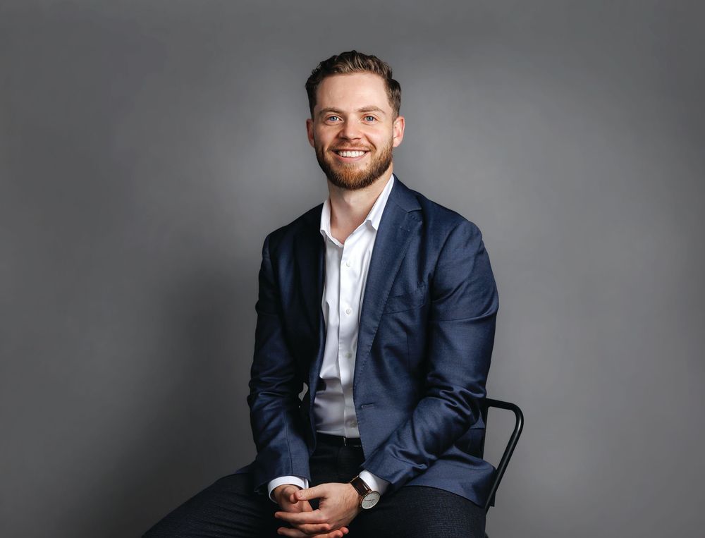 Professional headshot of someone in a navy suit and white shirt sitting casually against a gray studio background.