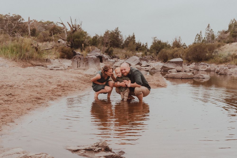 Three figures crouch down to explore the shallow water's edge along a rocky desert riverbank.