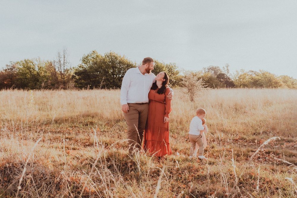 A family in a rustic field during golden hour, with tall dried grass and autumn trees in the background.
