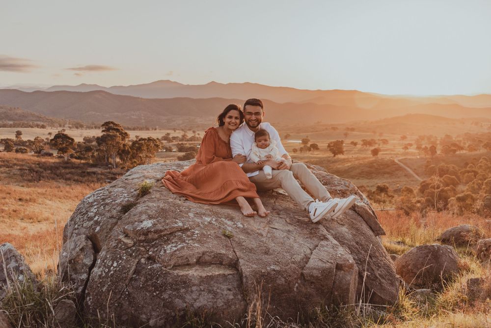 Two people sit together on a large rock formation at sunset in a mountainous landscape.