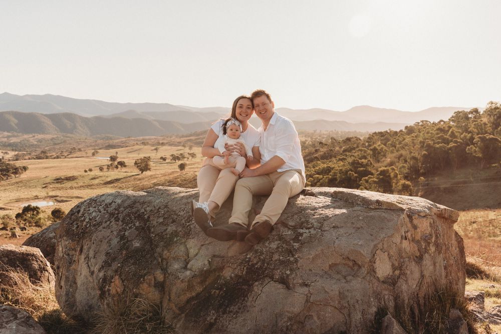 A couple sits together on a large boulder overlooking a golden valley landscape.