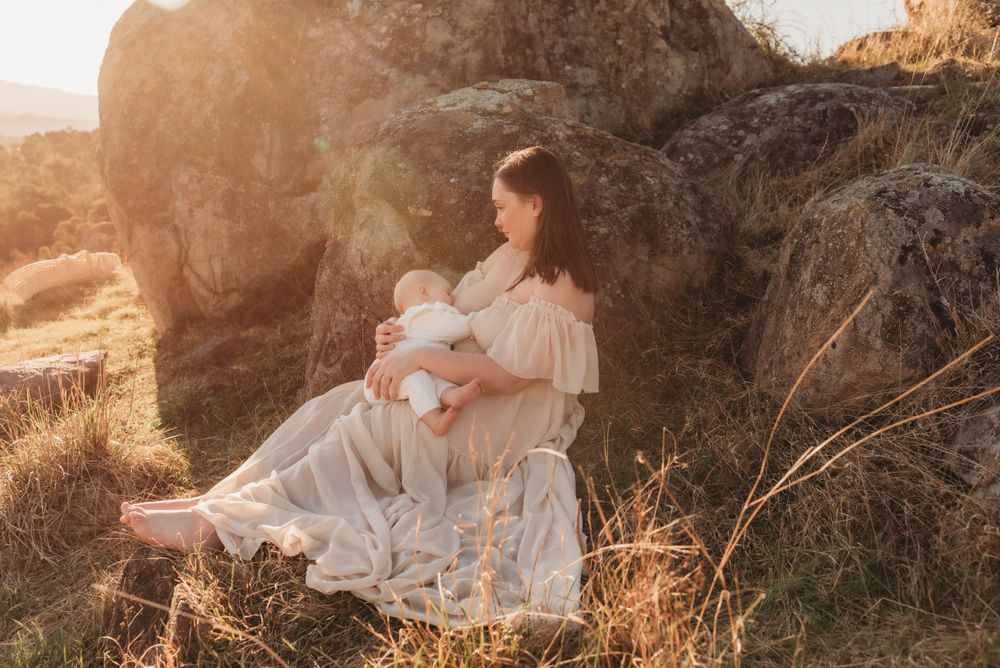 A person in a flowing white dress sits with a baby on a rocky outcrop at sunset.