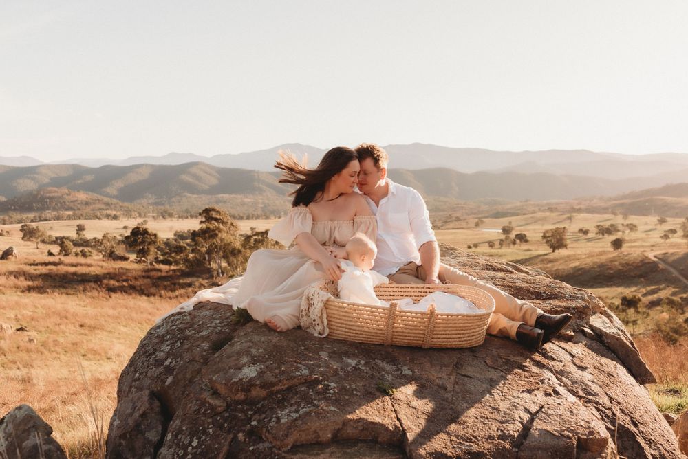 Two people share a picnic basket on a large rock overlooking rolling hills at sunset.