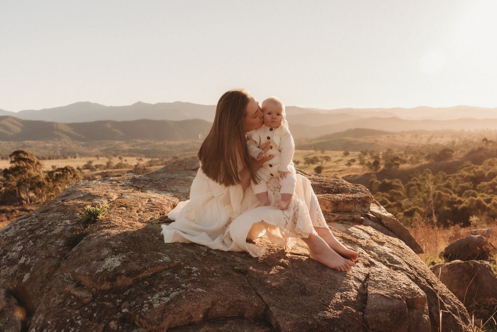 A woman in white holds a baby while sitting on a rocky outcrop during golden hour.