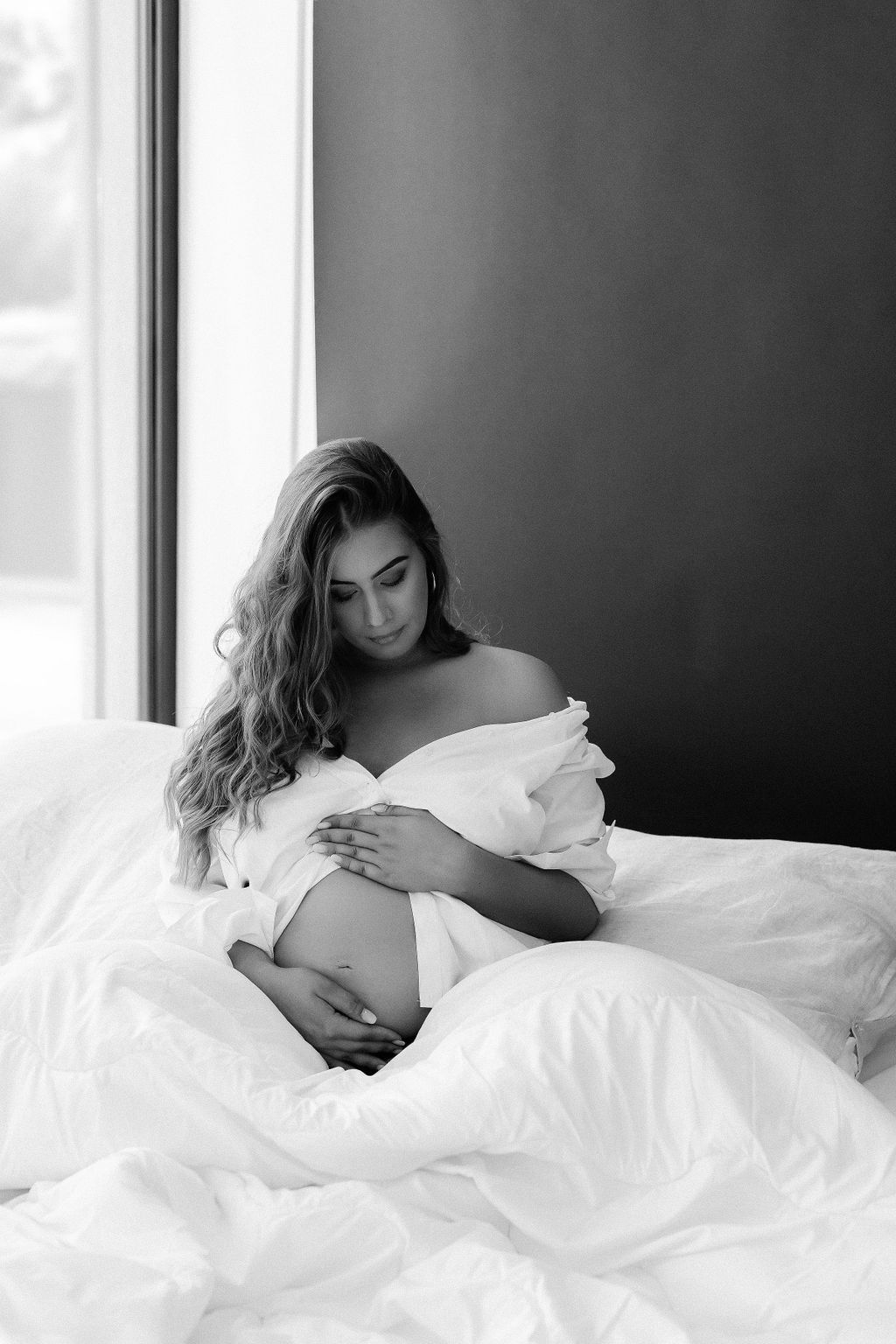 Čiernobiela tehotenská fotografia ženy sediacej na posteli pri okne, zahalenej v bielej prikrývke, s jemným prirodzeným svetlom. Monnaph