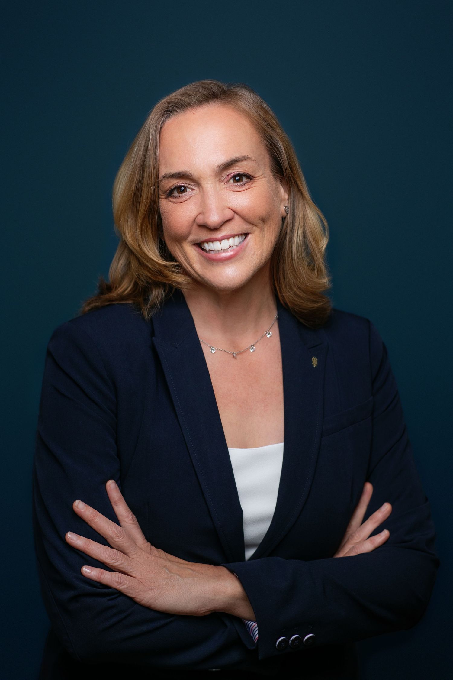 Professional headshot of a person wearing a navy blazer and white top smiling warmly against dark background.