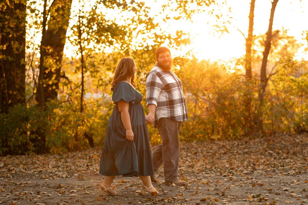 Warm, golden image taken by an engagement photographer of a couple holding hands and walking along a tree line in Twin Lakes, Iowa.