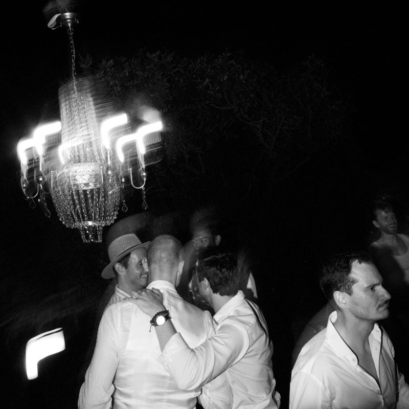 Black and white photo of men in white shirts at an evening party under a glowing crystal chandelier.
