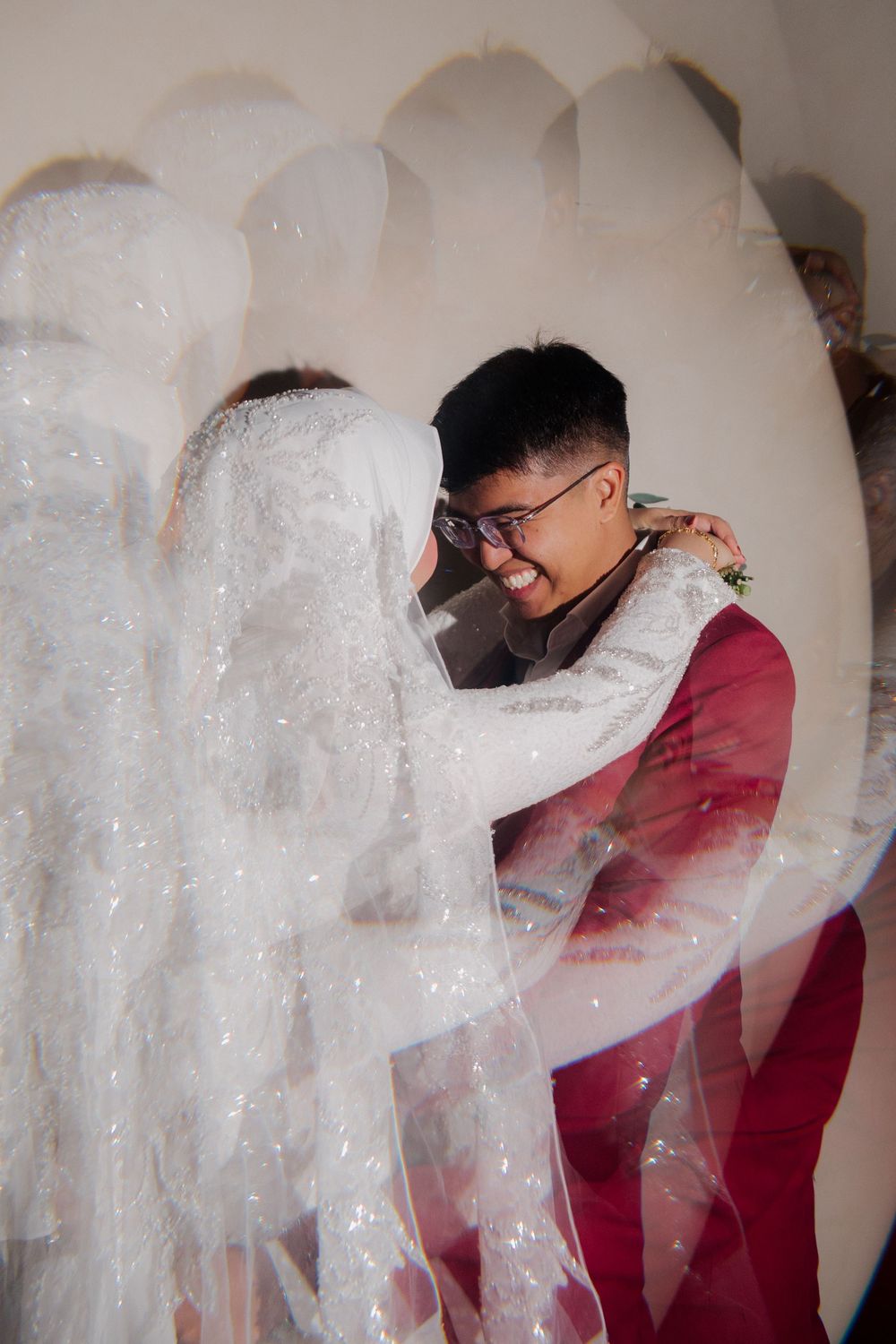A joyful groom in a red suit embraces his bride in a white lace wedding gown and veil, captured through a dreamy lens effect.
