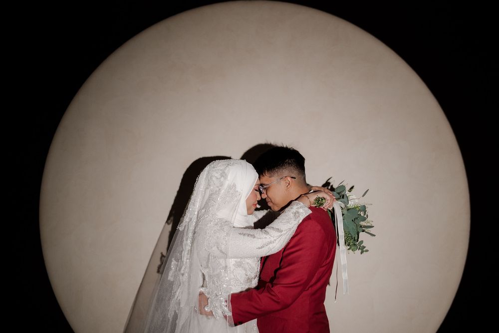A bride in white hijab and groom in red suit share a romantic moment holding a bouquet against a circular backdrop.