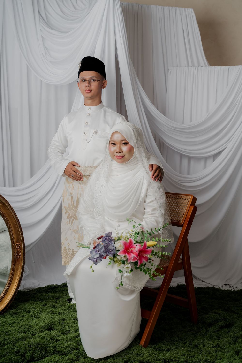 Muslim couple in white traditional wedding attire, bride seated holding pink flowers, groom standing behind, white draped backdrop.