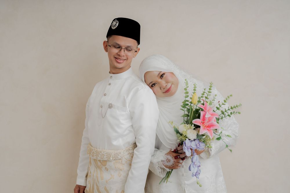 Muslim couple in white traditional attire, groom in baju melayu and bride holding colorful bouquet, posing for wedding photo.