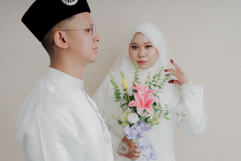Muslim couple in white attire, groom in songkok hat, bride holding pink lily bouquet during traditional Malay wedding ceremony.