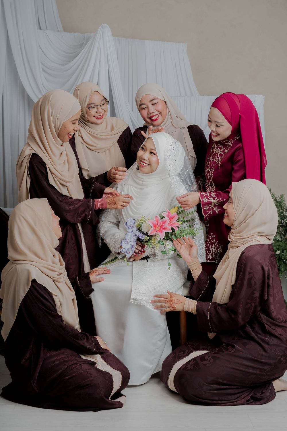 Muslim bride in white hijab surrounded by bridesmaids in dark maroon and beige hijabs holding floral bouquet.