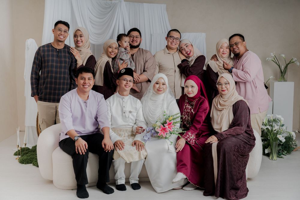 A Muslim wedding group photo with bride and groom in white seated among elegantly dressed family members.