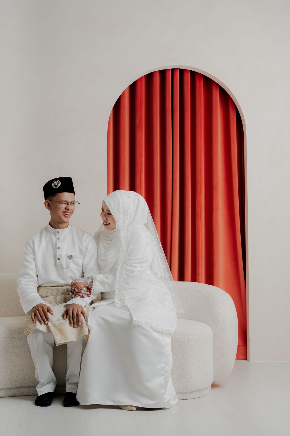 Malay couple in white traditional wedding attire sitting together in front of a red arched curtain backdrop.