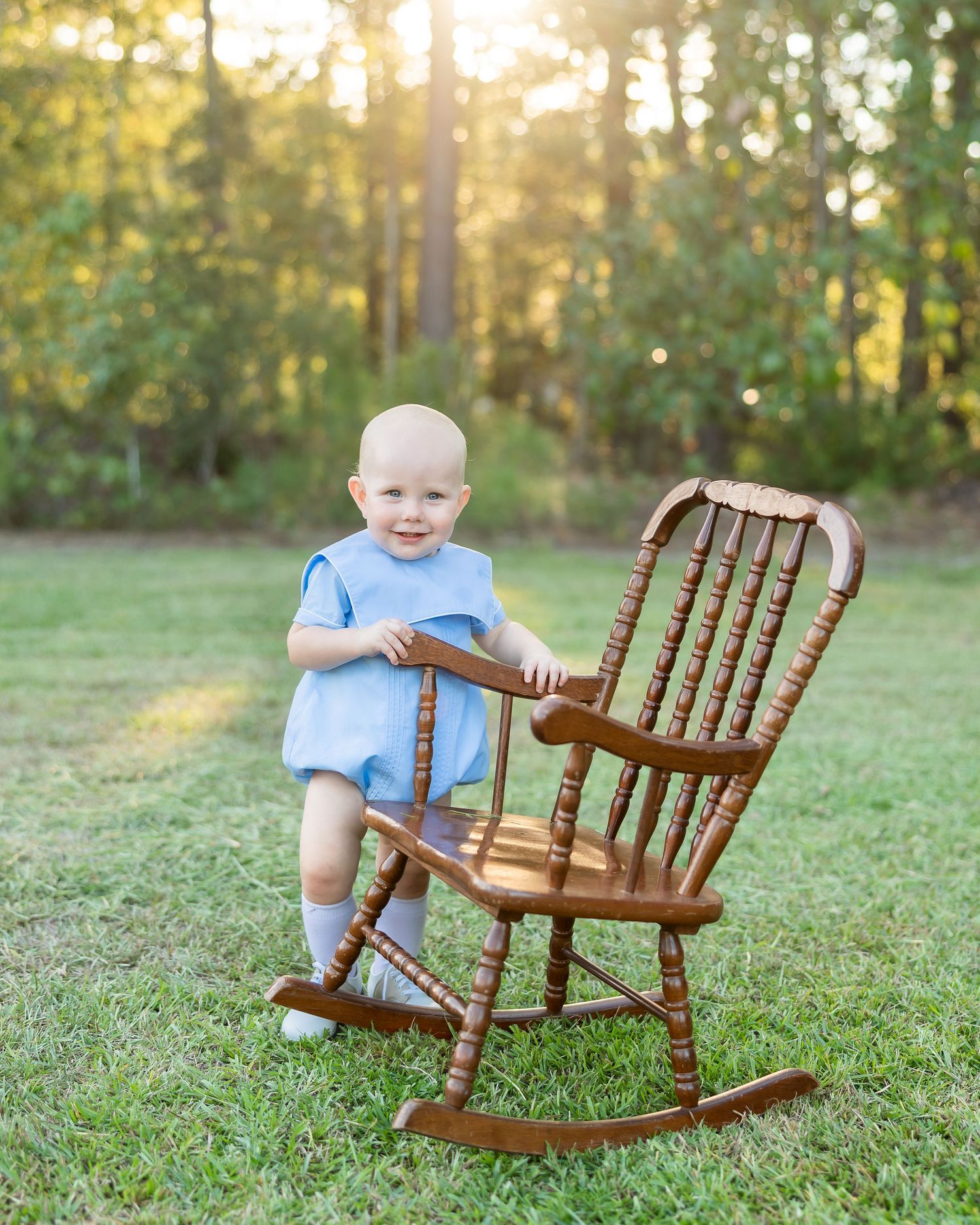 Smiling baby in blue romper standing beside a wooden rocking chair on green grass with sunlit trees.