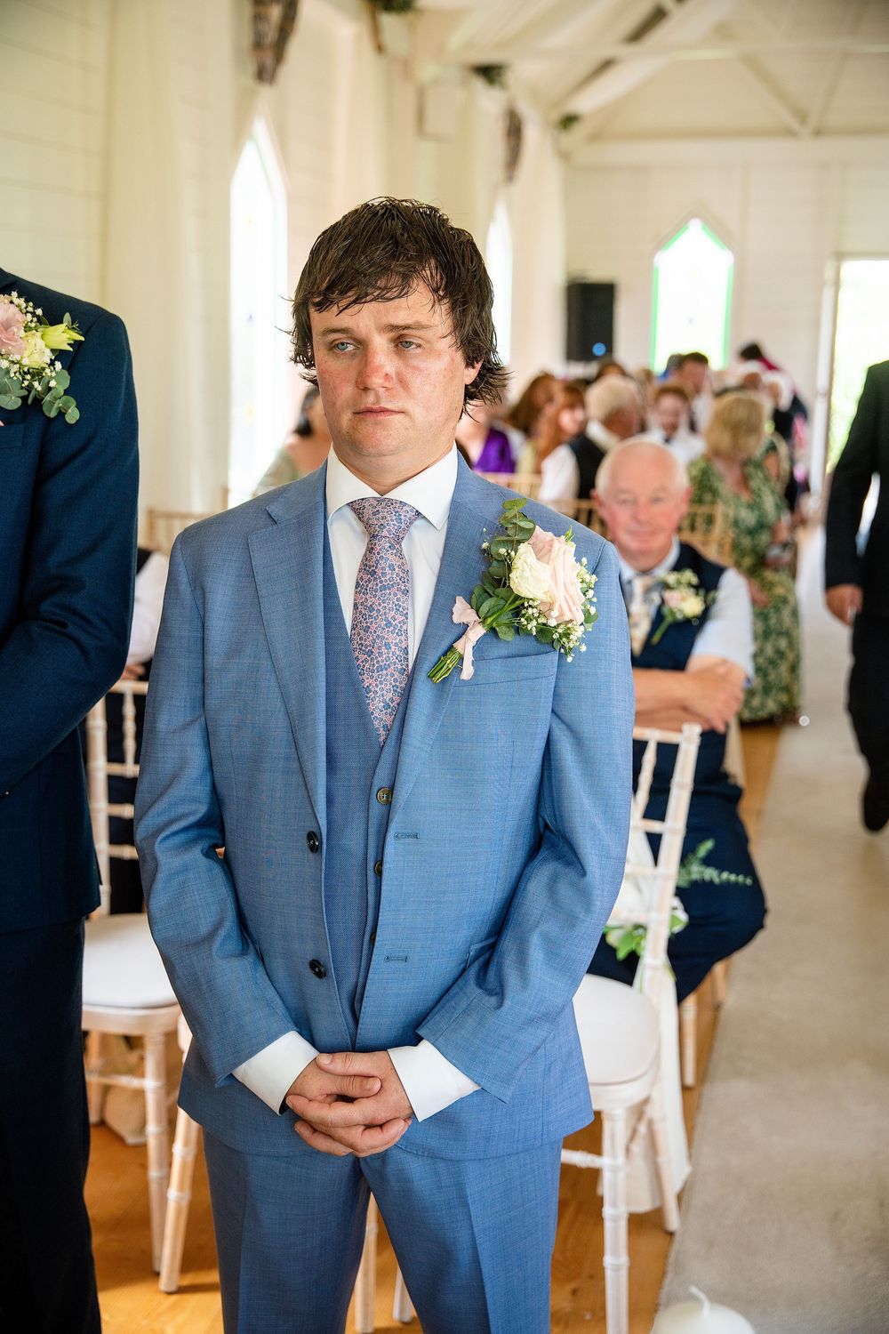 Groom in light blue three-piece suit with floral boutonniere stands at wedding altar awaiting bride.
